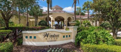 A landscaped entrance with a sign for Windsor Hills Clubhouse, surrounded by palm trees and well-maintained hedges.