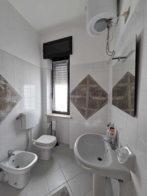 A bathroom with a toilet, bidet, sink, and a window with lace curtains.