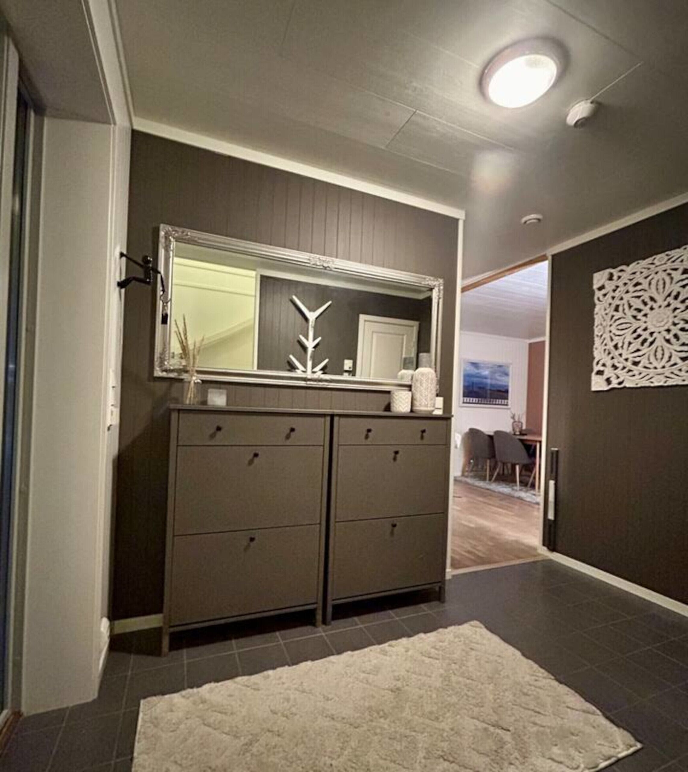 A hallway with a large mirror, a dark brown wardrobe, a white rug, and a decorative wall piece.
