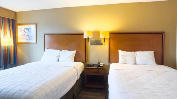 Quarto de hotel com duas camas, mesa de cabeceira com abajur e quadro pendurado na parede.