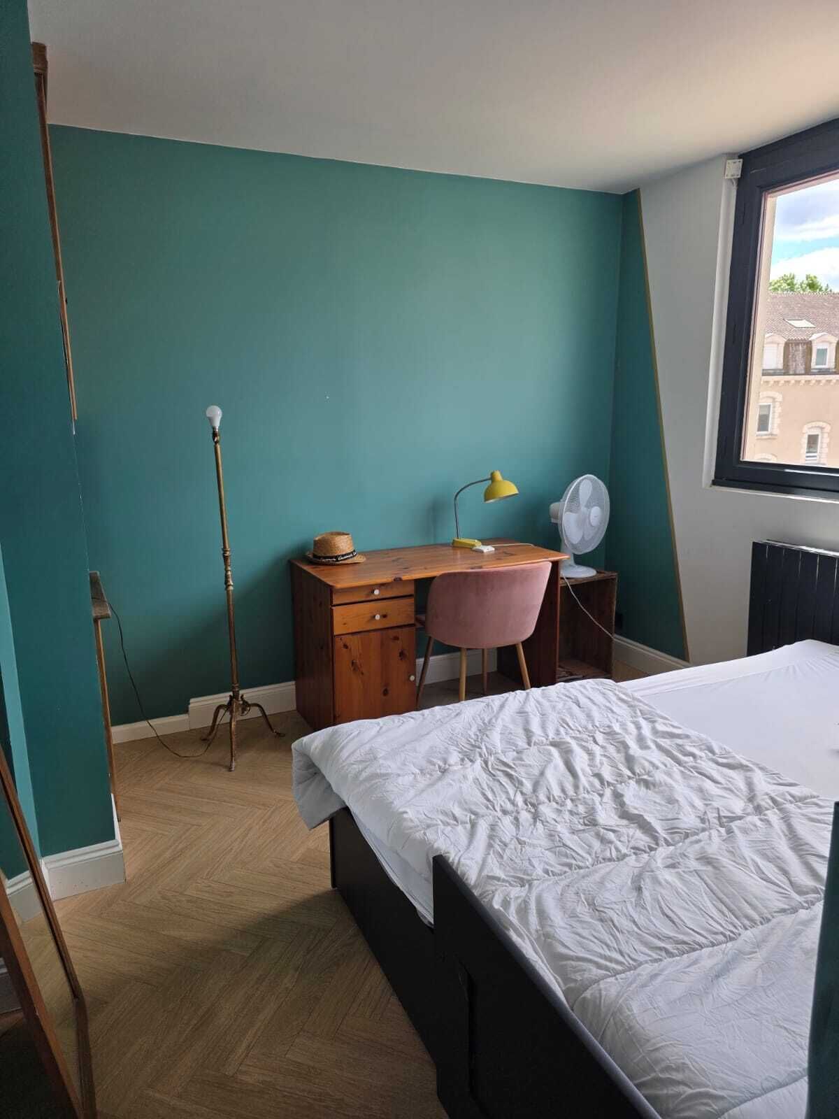A bedroom with teal walls, a wooden desk, a pink chair, a bed with white bedding, a standing lamp, and a window with a view.
