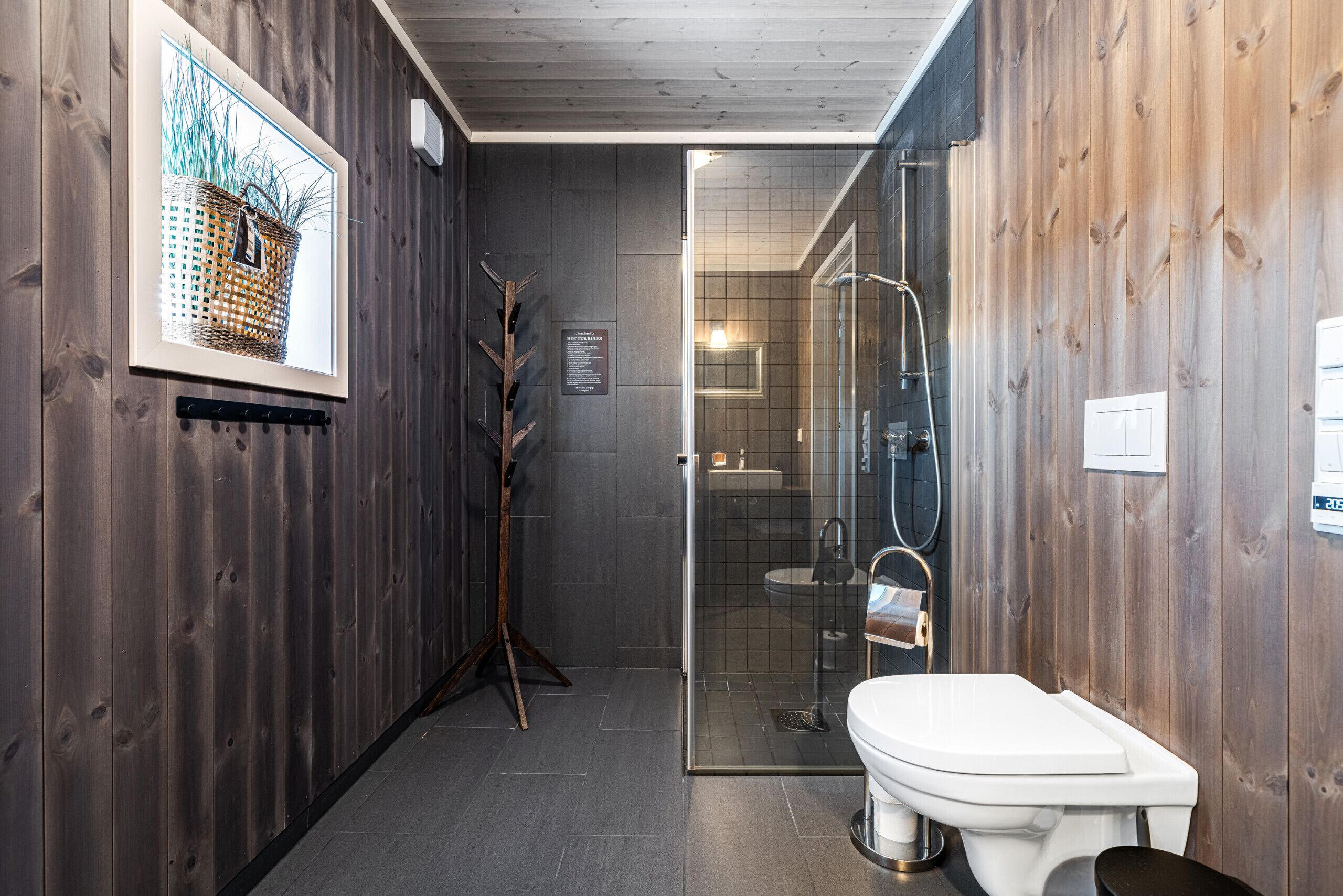 Een moderne badkamer met een houten wand, een glazen douchecabine, een wit toilet en een lijst met een afbeelding van een mand.