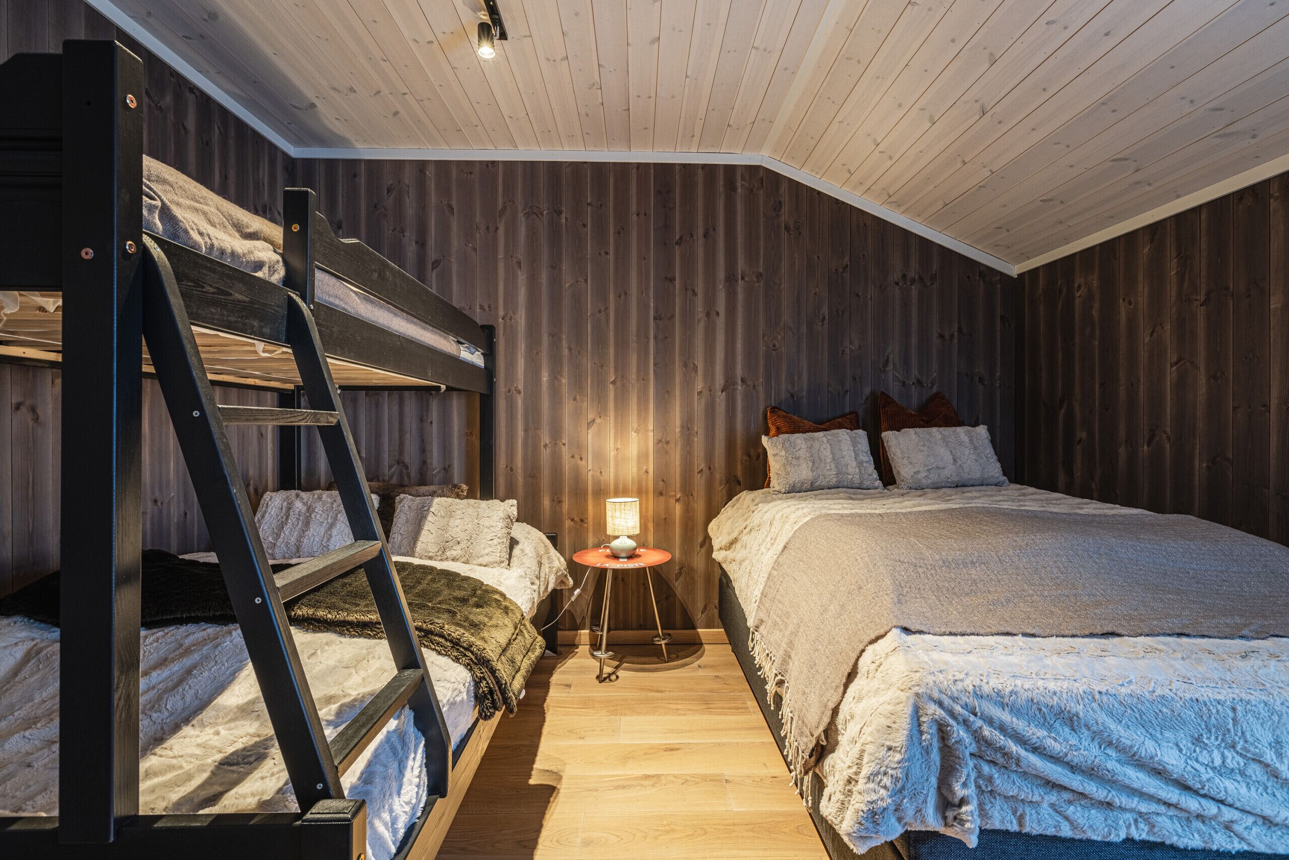 Une chambre avec un lit superposé, un plafond et des murs en bois, une petite table ronde avec une lampe, et une table de chevet avec une lampe.