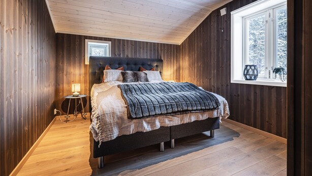 Ein Schlafzimmer mit Holzbalkendecke, einem Bett mit dunklem Kopfteil, einem Nachttisch mit Lampe und einem Fenster mit Blick auf Bäume.