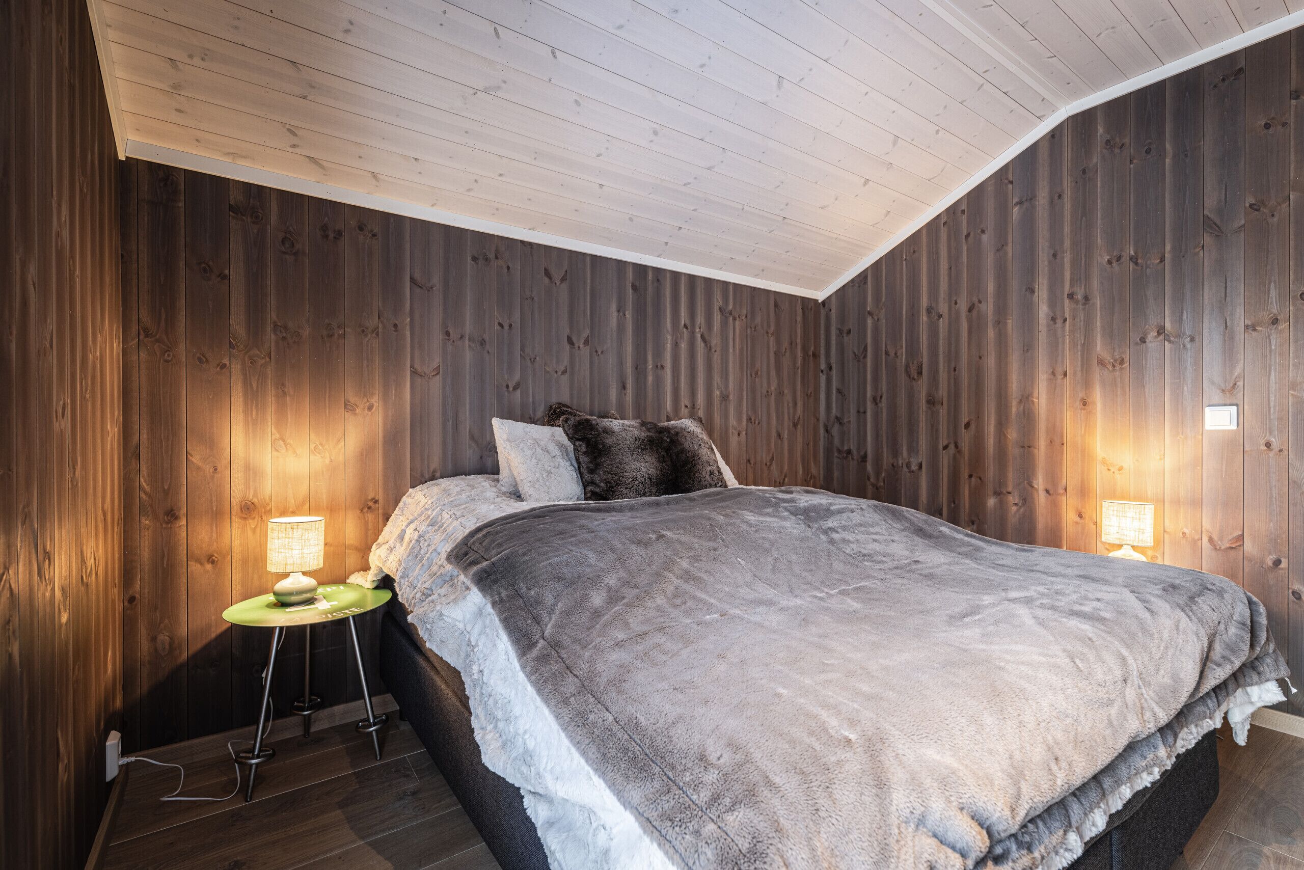 A bedroom with a wooden wall, a bed with a grey blanket, and two bedside lamps.