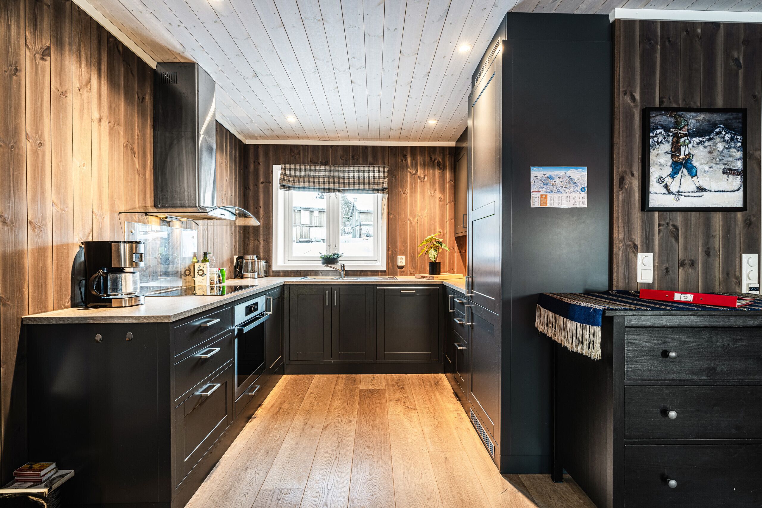 Une cuisine moderne avec des meubles de couleur sombre, un plancher en bois, une grande fenêtre et un tableau accroché au mur.