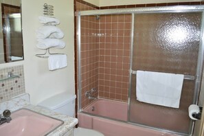 Un baño con una bañera rosa, ducha con mampara de vidrio y toallero con toallas dobladas.