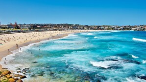 Una playa costera con aguas cristalinas, una orilla arenosa y edificios al fondo.