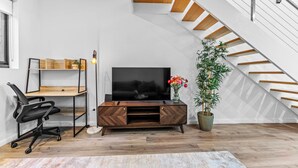 Una sala moderna con unidad de entretenimiento de madera, una silla de oficina negra, un televisor de pantalla plana grande, una planta en maceta y una escalera.