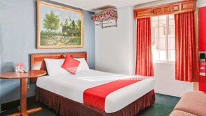 Hotellihuone, jossa on sänky, pieni pöytä, seinällä taulu ja ikkuna, jossa on verhot.