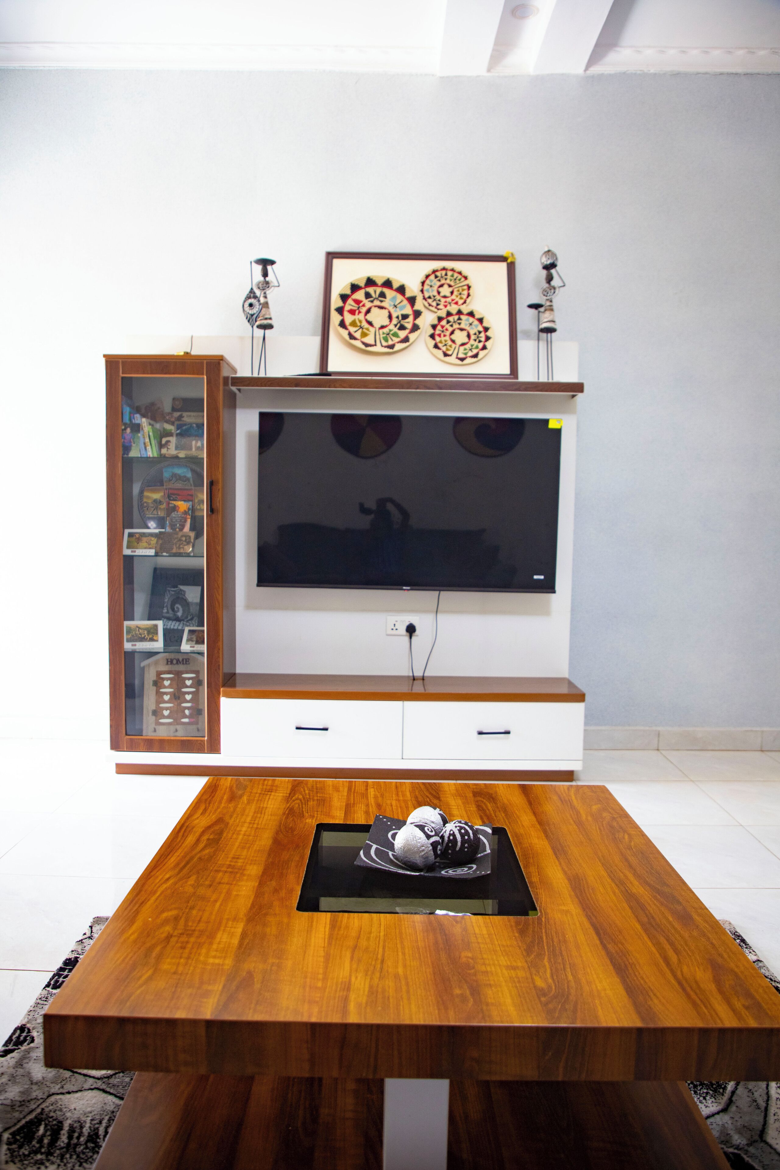Un salon moderne avec une table basse en bois, un téléviseur à écran plat fixé sur un meuble de télévision blanc, et un meuble vitré contenant des objets décoratifs.