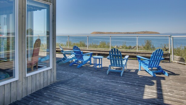 A wooden deck with blue Adirondack chairs and a glass wall overlooking a body of water. - Direct Beach Access! Quiet Diamond Point Retreat (Sequim)