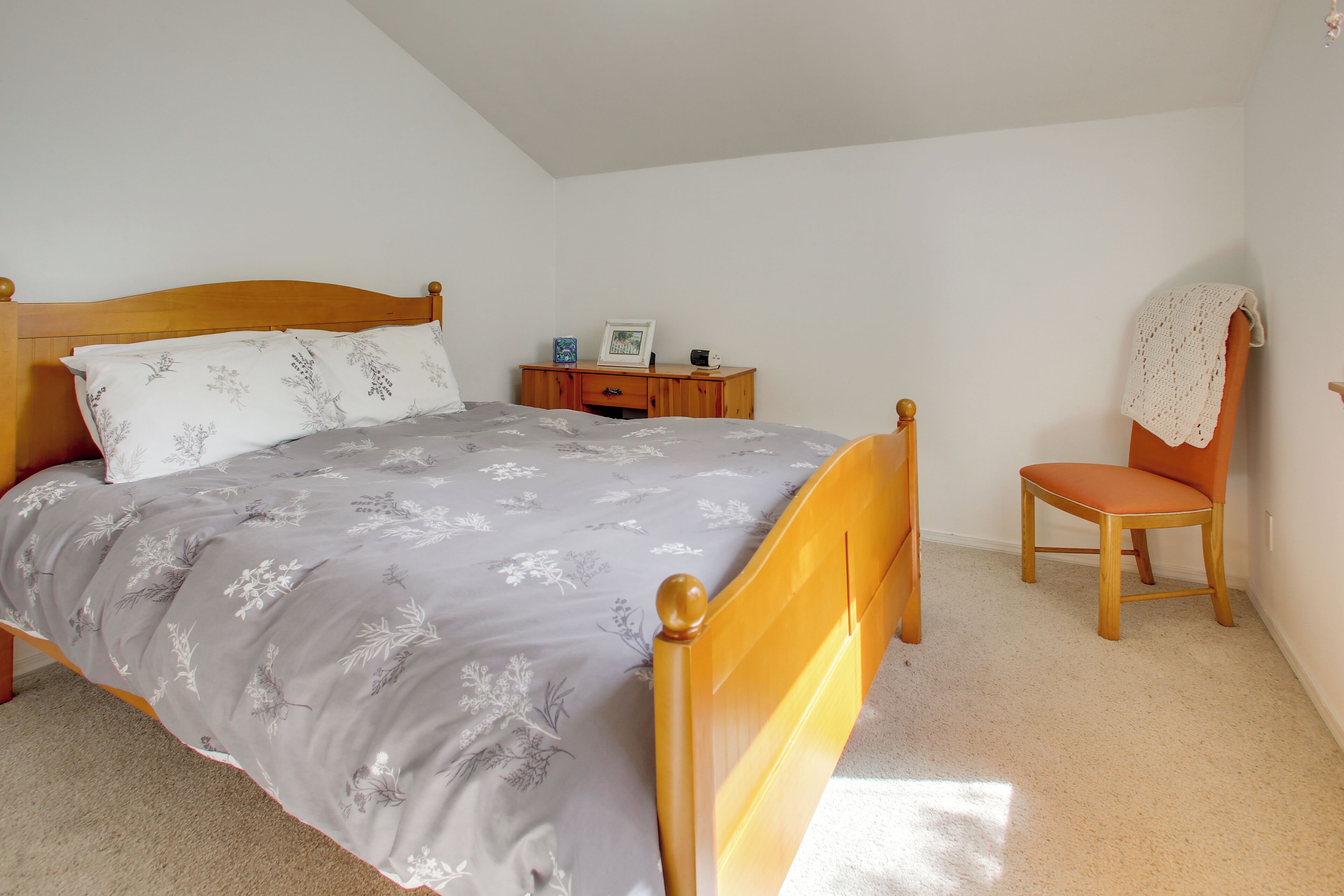 Un dormitorio con una cama, una mesita de noche de madera, una silla y una pequeña mesa con un marco de fotos.