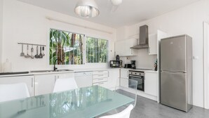 A modern kitchen with white cabinetry, a stainless steel refrigerator, a glass dining table, and a window with a view of greenery. - Magnífica Villa 5 dormitorios con piscina privada cerca Banus (Marbella)