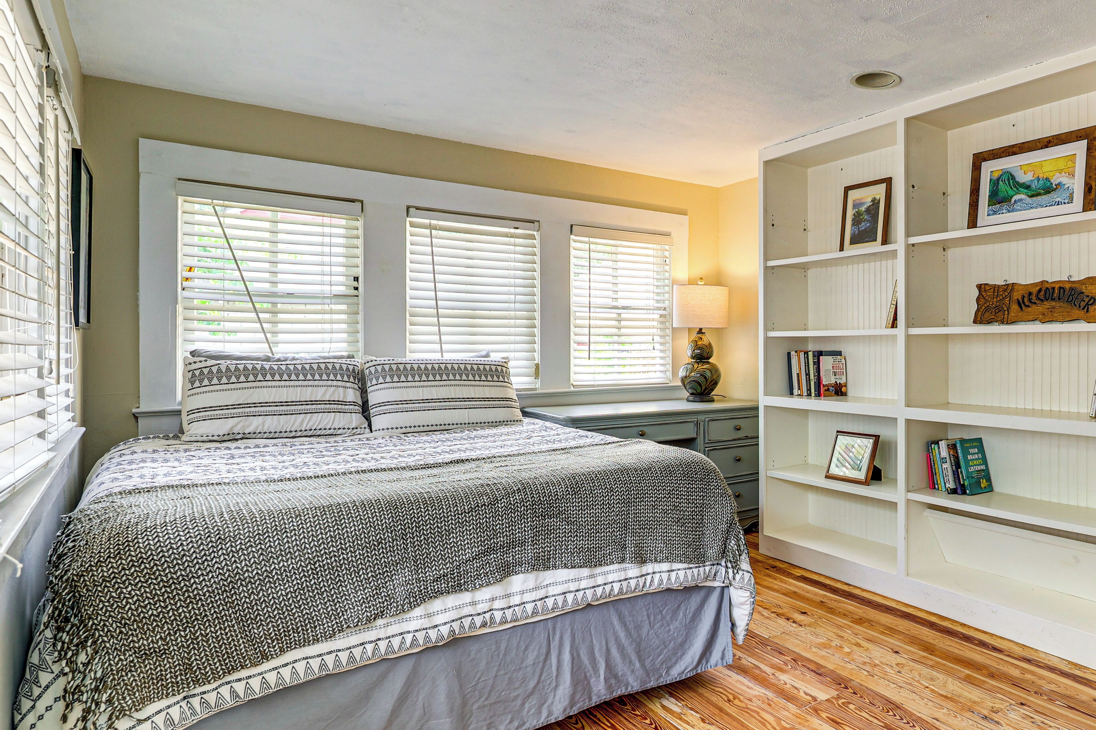 A bedroom with a bed, two windows with blinds, a bedside table with a lamp, and a bookshelf.