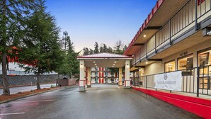 L’entrée d’un hôtel, avec un espace de stationnement couvert, une façade de couleur rouge et beige, et une banderole indiquant le nom de l’hôtel.