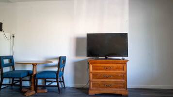 A room with a TV on a wooden dresser, two blue chairs around a small round table, and a plain white wall.
