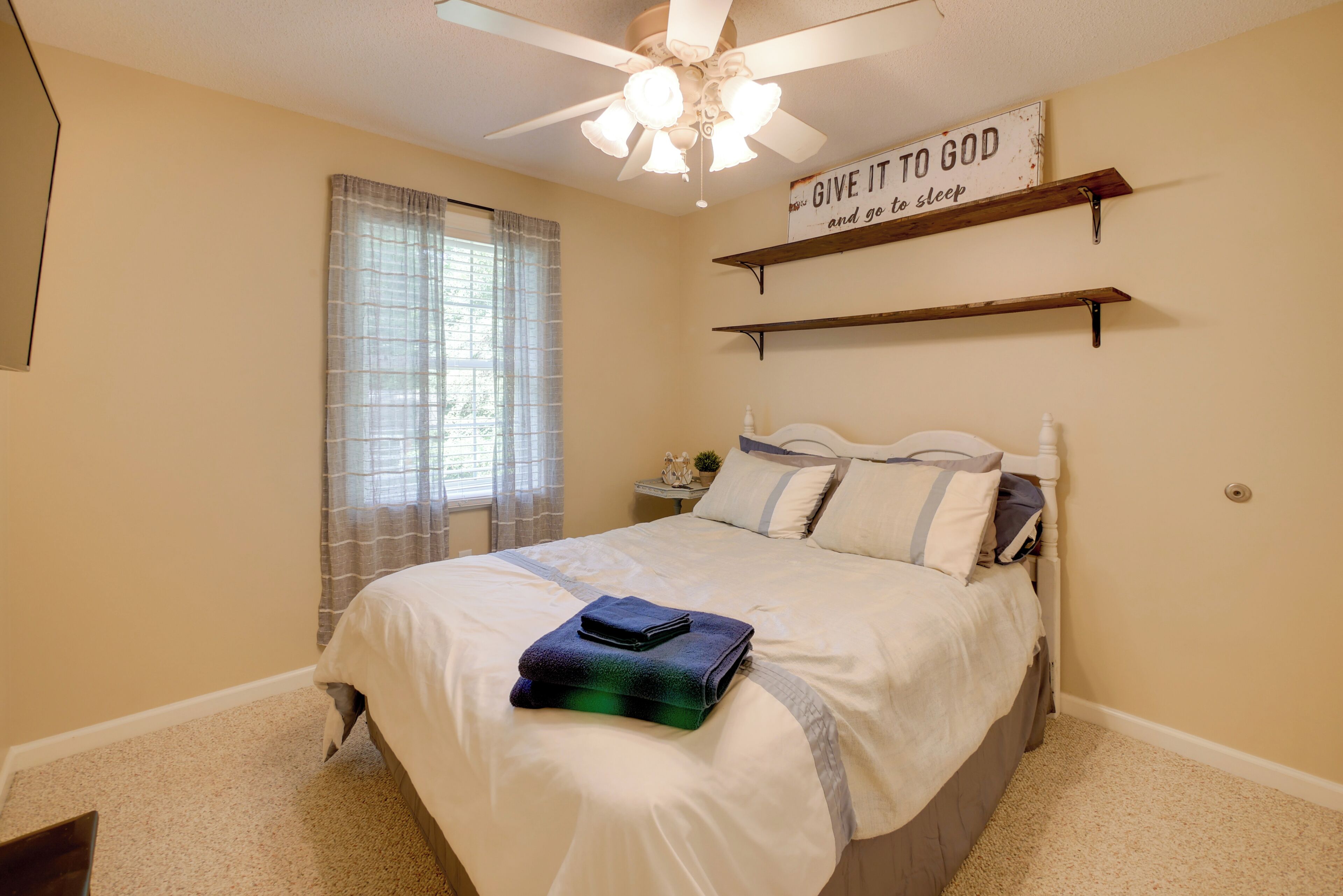 Quarto com cama, ventilador de teto, janela com cortinas e uma frase motivacional na parede.