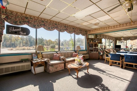 A room with a sofa, a coffee table, a dining table with chairs, a window with a 'Thank You' sign, and a view of trees.