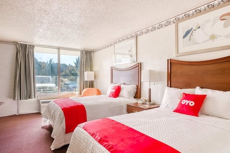 A hotel room with two beds, a window with a view of parked cars, a wooden headboard, and a framed picture on the wall.