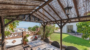A covered outdoor seating area with a table and chairs, a thatched roof, and a view of a white building and greenery.