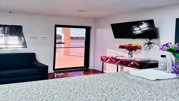Una camera d'albergo con TV a schermo piatto, una scrivania con un vaso di fiori, un tavolo rosso e una porta scorrevole in vetro che dà su un balcone.