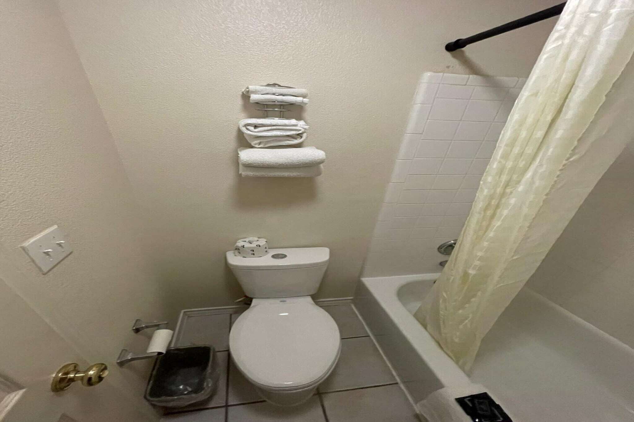 A small bathroom with a toilet, a towel rack with folded towels, a trash bin, and a bathtub with a shower curtain.