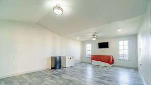 A spacious room with a high ceiling, a ceiling fan, a flat-screen TV mounted on the wall, and a window providing natural light.