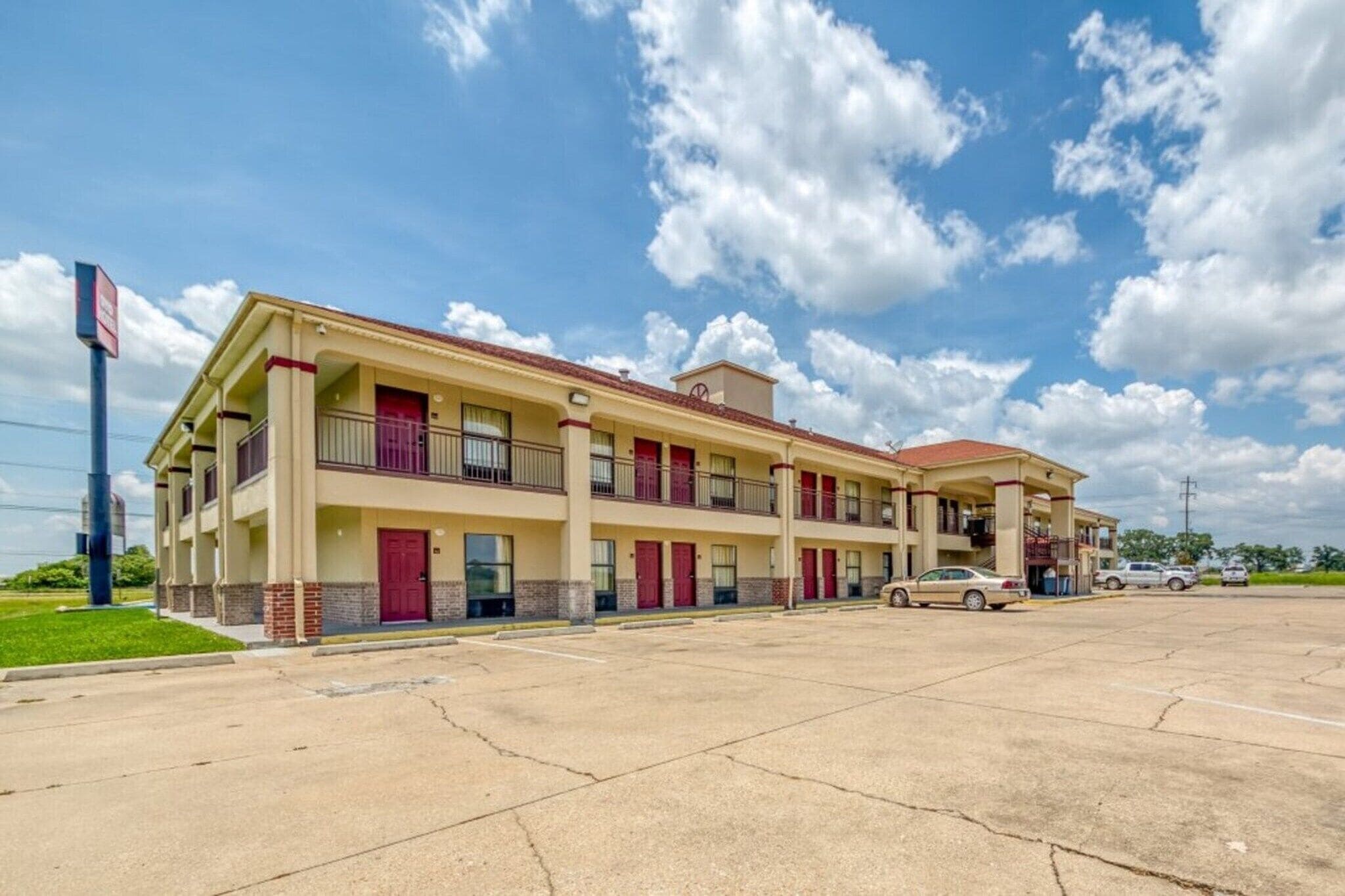 Un motel à plusieurs étages, aux couleurs rouge et beige, avec un parking et un ciel dégagé.