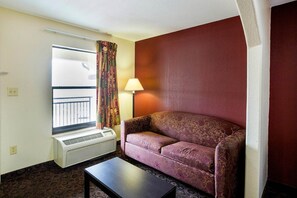 A hotel room with a sofa, a table, a window with curtains, a lamp, and an air conditioning unit. - 2 Queen Bed | Port Allen LA (Port Allen LA)