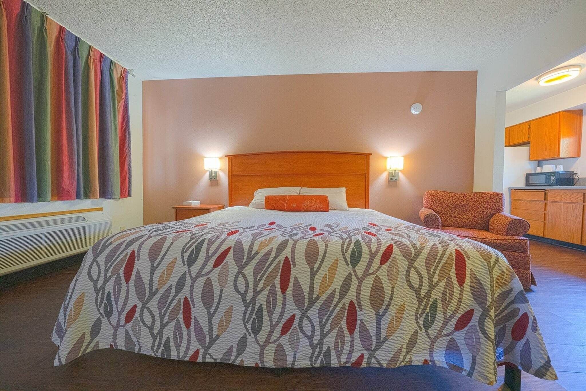 A hotel room with a bed, a chair, a nightstand, and a wall-mounted heater.