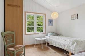 A room with a wooden chair, a small white table, a bed with a white blanket, a window with a view of trees, and a hanging light fixture. - Holiday Close to Forest, Fjord and Beach (Kulhuse)