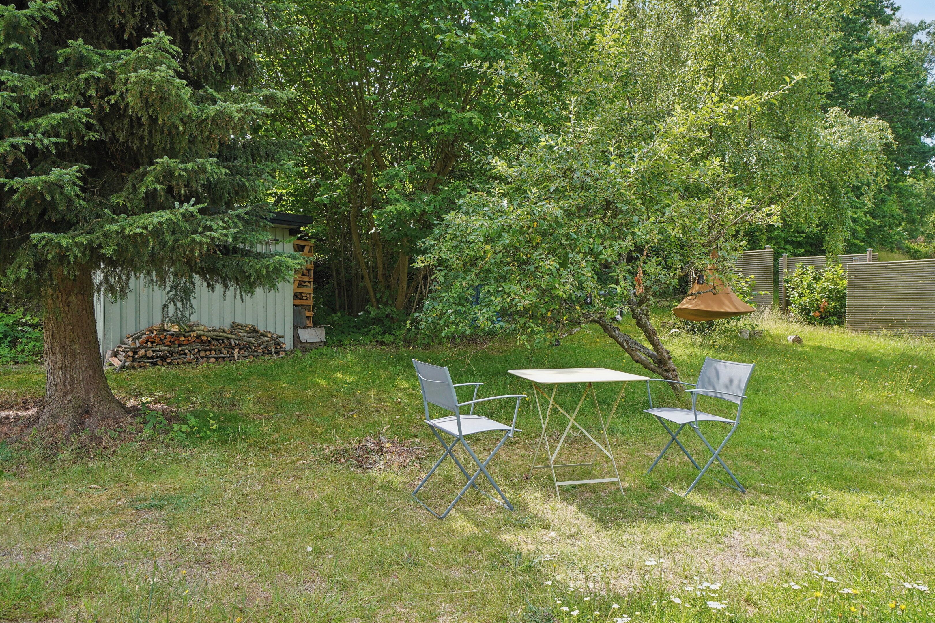 An outdoor seating area with a table and two chairs, surrounded by trees and a pile of logs.