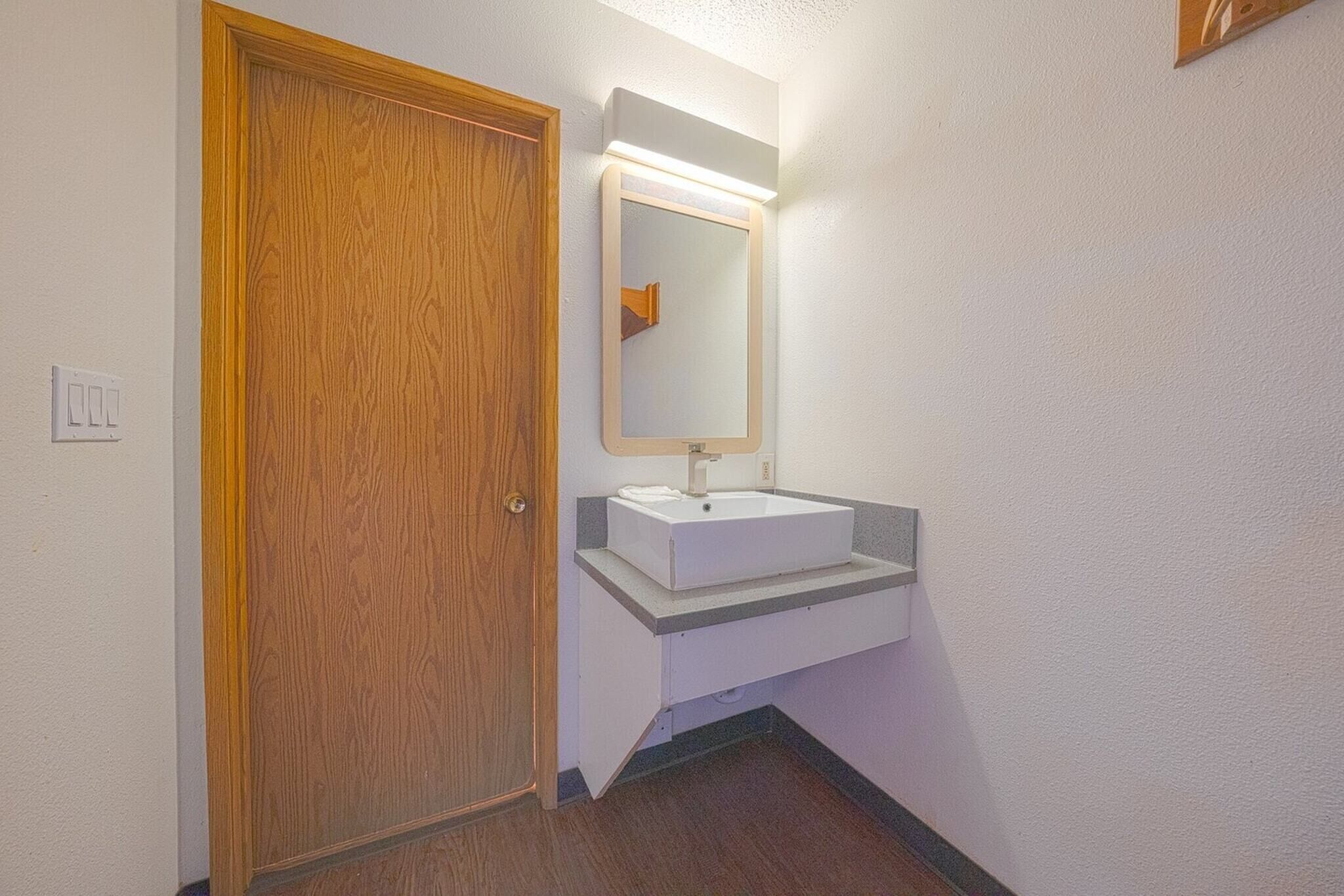 Un baño con puerta de madera, un lavabo blanco y un espejo con luz superior.
