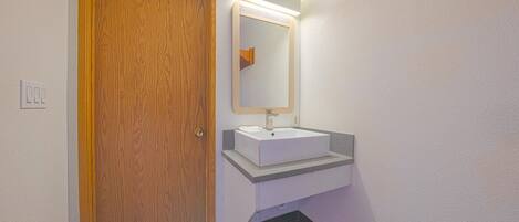 Un baño con puerta de madera, un lavabo blanco y un espejo con luz superior.