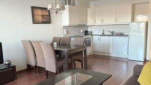 Una sala moderna con una mesa de centro de vidrio, un sofá con un cojín amarillo, un comedor con sillas y una cocina con gabinetes y electrodomésticos blancos.