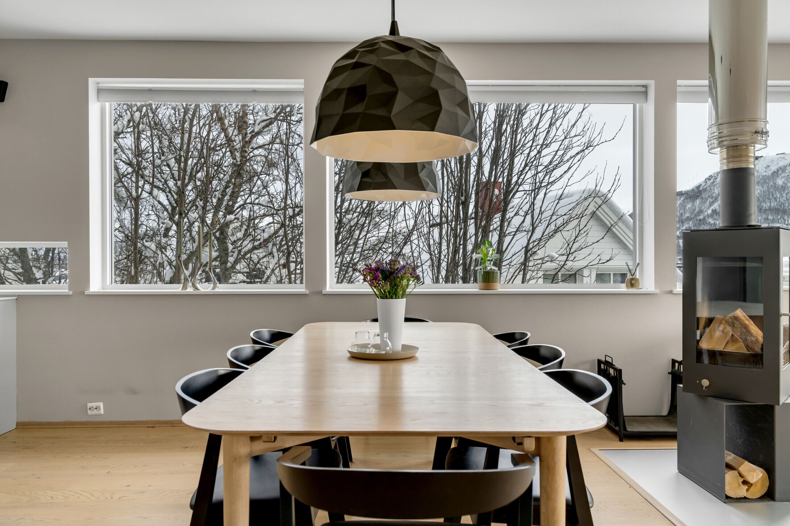 Een moderne eetruimte met een houten tafel, zwarte stoelen, een geometrische hanglamp en uitzicht op met sneeuw bedekte bomen door grote ramen.