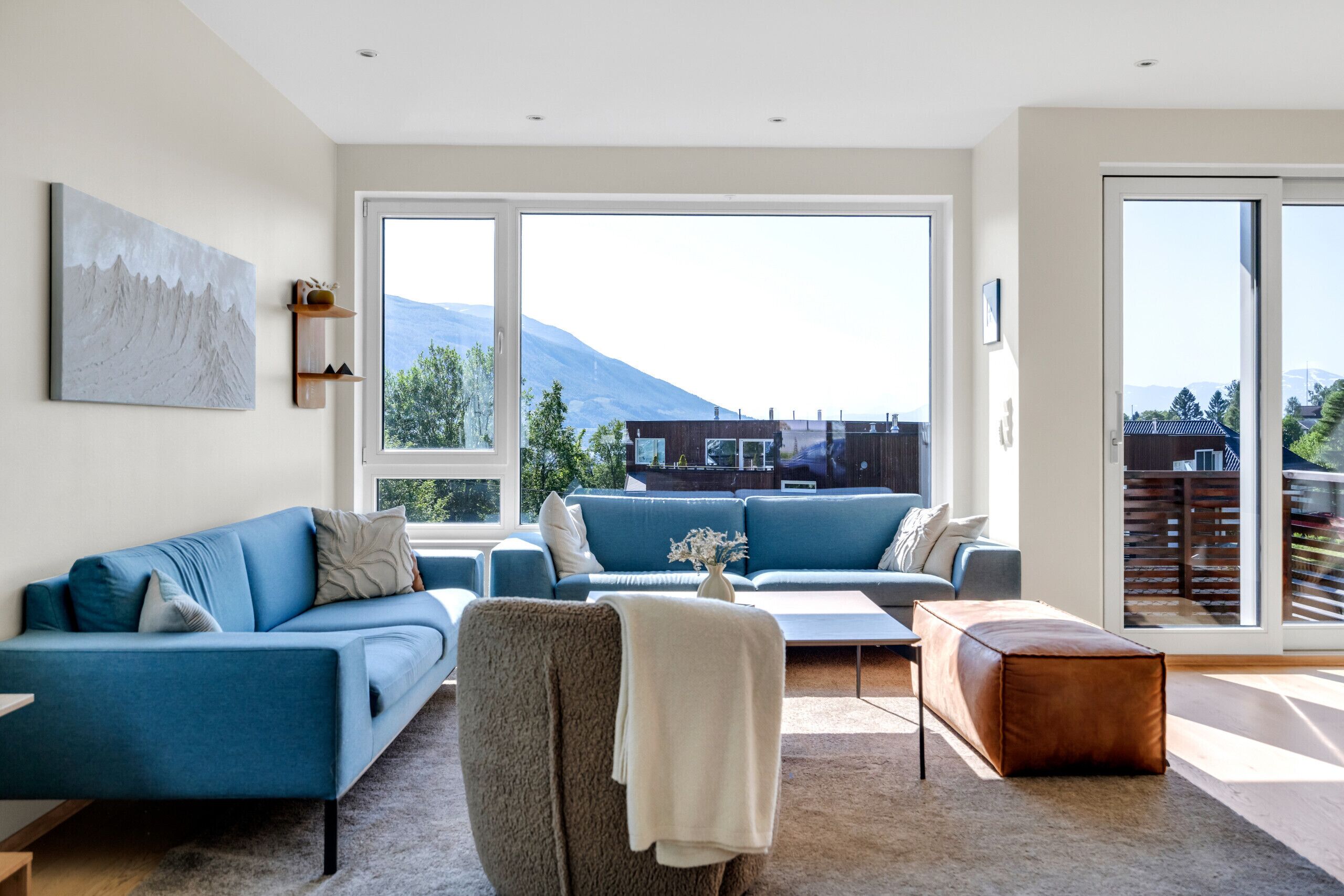 Un salon moderne avec un canapé bleu, un fauteuil marron et une table basse. De grandes fenêtres offrent une vue sur une montagne et un immeuble.
