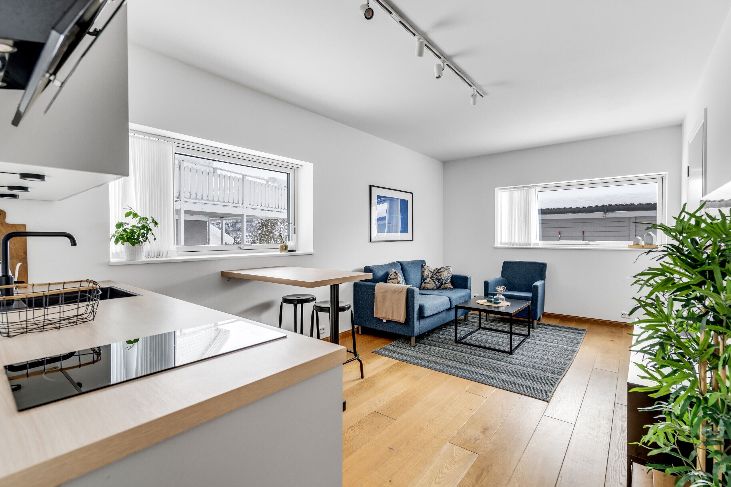 A modern kitchen with a countertop and sink, a dining area with a table and chairs, a blue sofa, and a view of the outside through large windows.