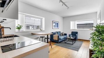 A modern kitchen with a countertop and sink, a dining area with a table and chairs, a blue sofa, and a view of the outside through large windows.