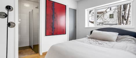 Un dormitorio con una cama, un ventanal, un cuadro abstracto rojo y una puerta que da a un baño.