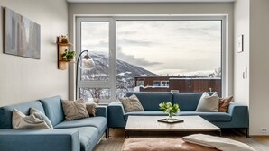 A modern living room with a blue sofa, a coffee table, and a view of mountains through large windows. - Northern Luxe Retreat (Tromsø)