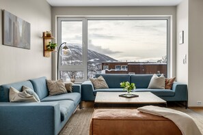 Un saló modern amb un sofà blau, una taula de centre i vista a les muntanyes a través de grans finestres.