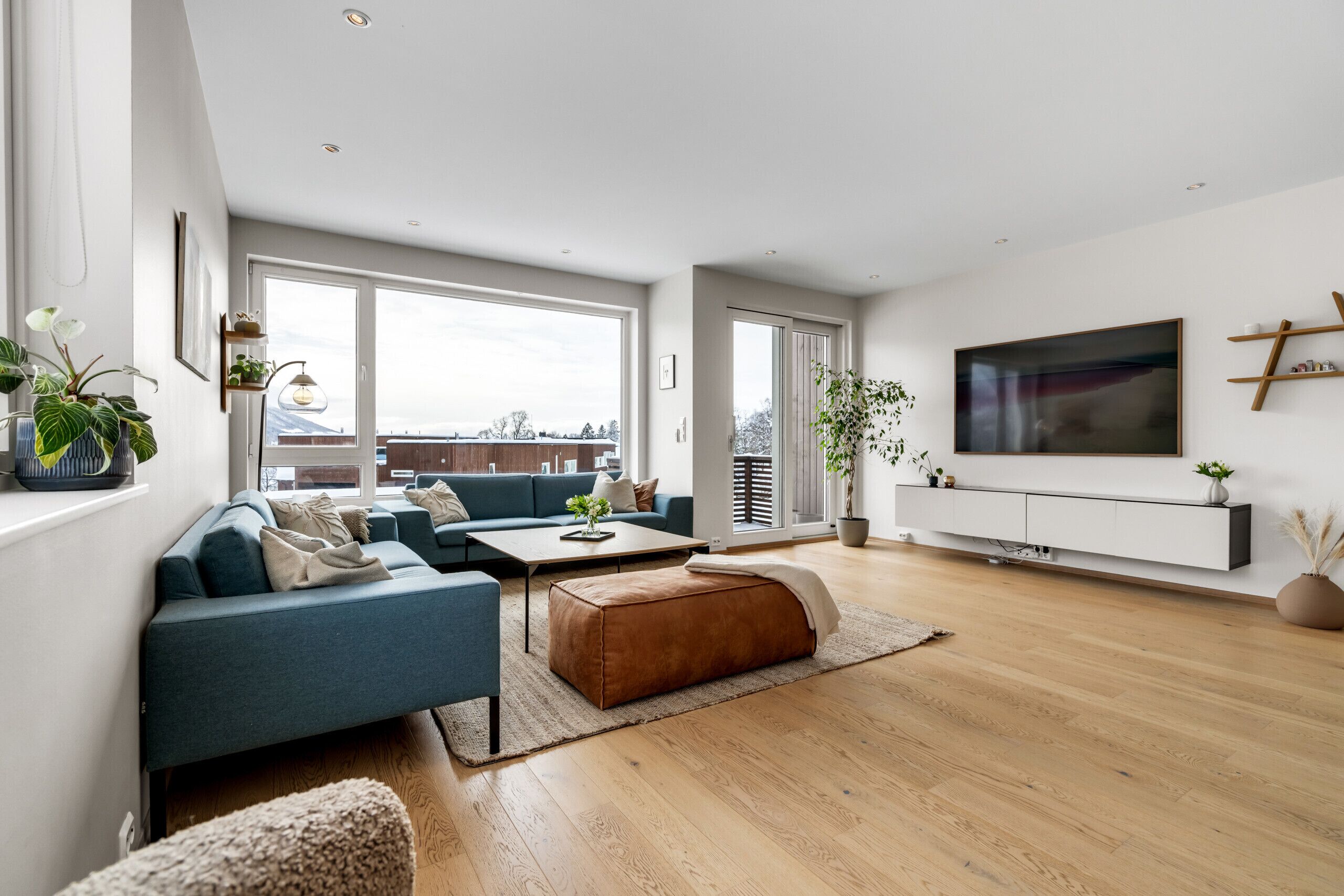 Una sala de estar moderna con sofá, mesa de centro y reposapiés, ventanas amplias y un televisor de pantalla plana montado en la pared.