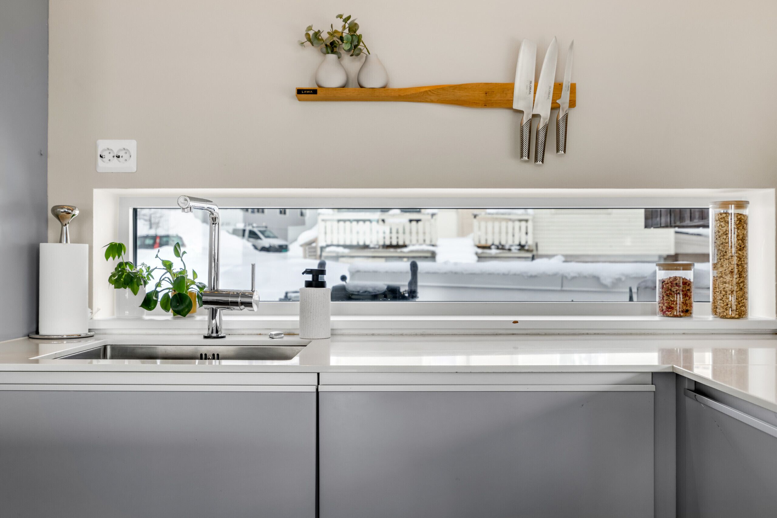 Una cocina con ventana, un lavamanos, un grifo, una planta y una estantería con frascos y utensilios.