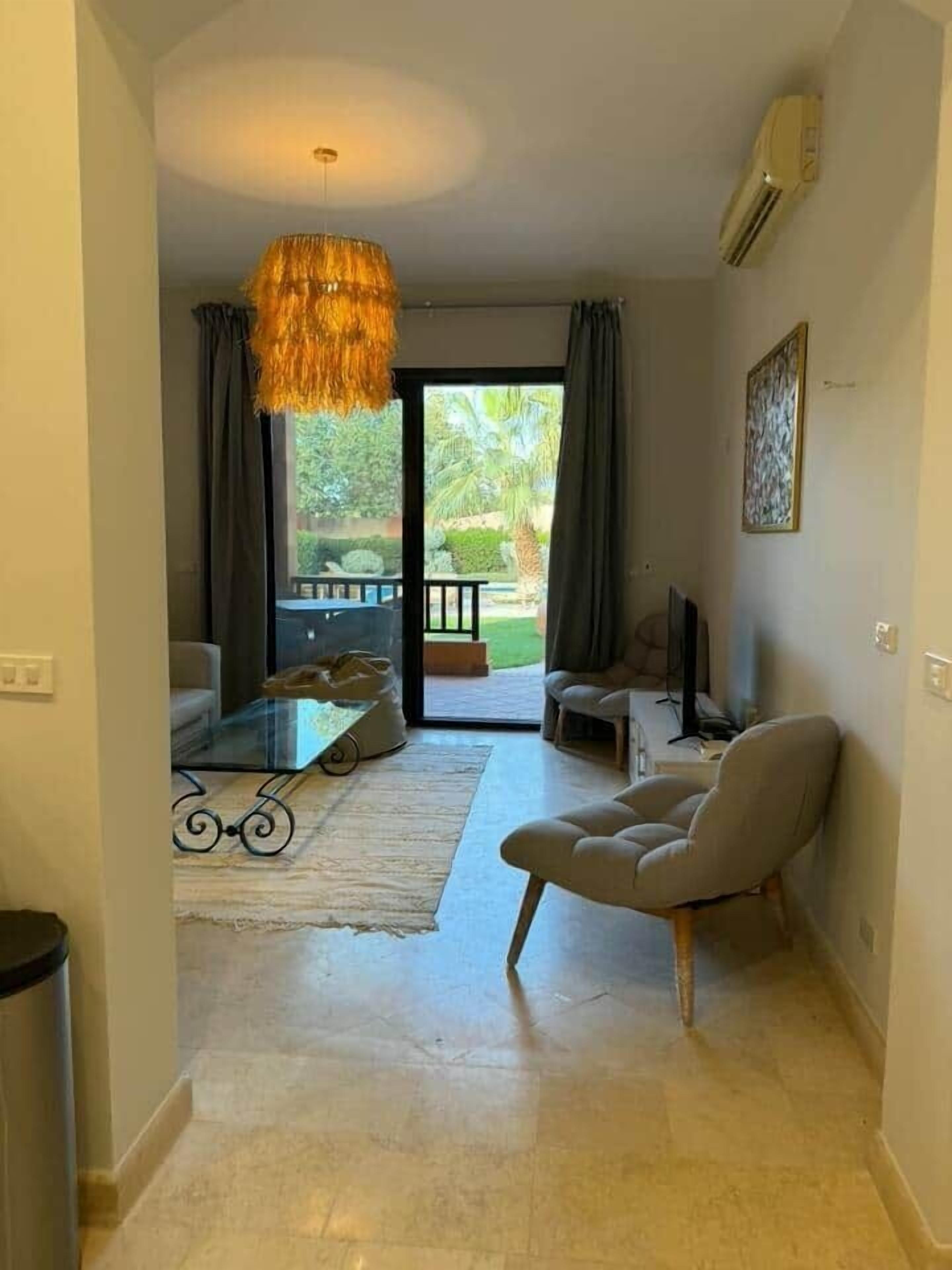 A living room with a glass coffee table, a sofa, a chair, a hanging light fixture, and a view through a glass door to an outdoor area.