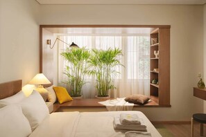 Habitación de hotel con cama, lámparas de noche, ventana con plantas, estante con libros y mesita de noche con toallas.