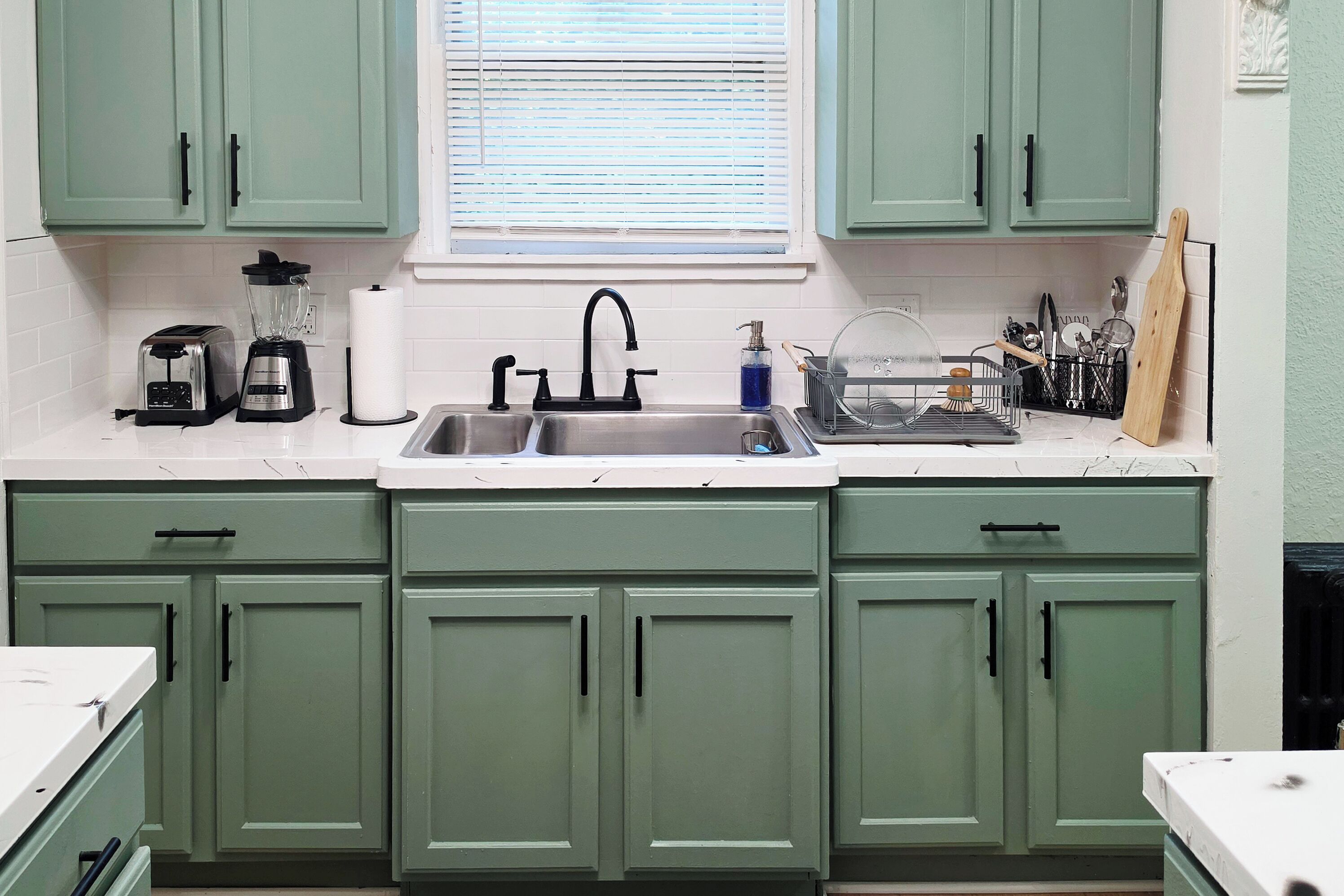 A kitchen with green cabinets, a double sink, a toaster, a blender, and a window with blinds.
