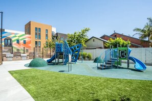 A playground with slides and climbing structures, surrounded by a grassy area and buildings in the background.
