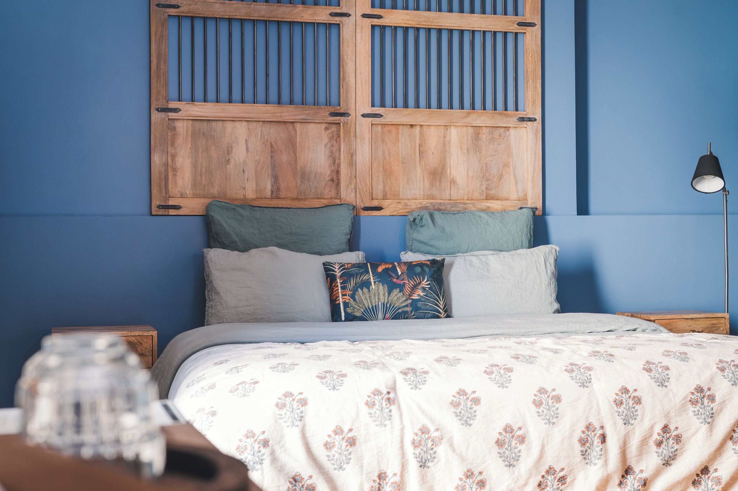 A bedroom with a wooden headboard, a bed with floral bedding, two pillows, a bedside lamp, and a nightstand with a glass jar.